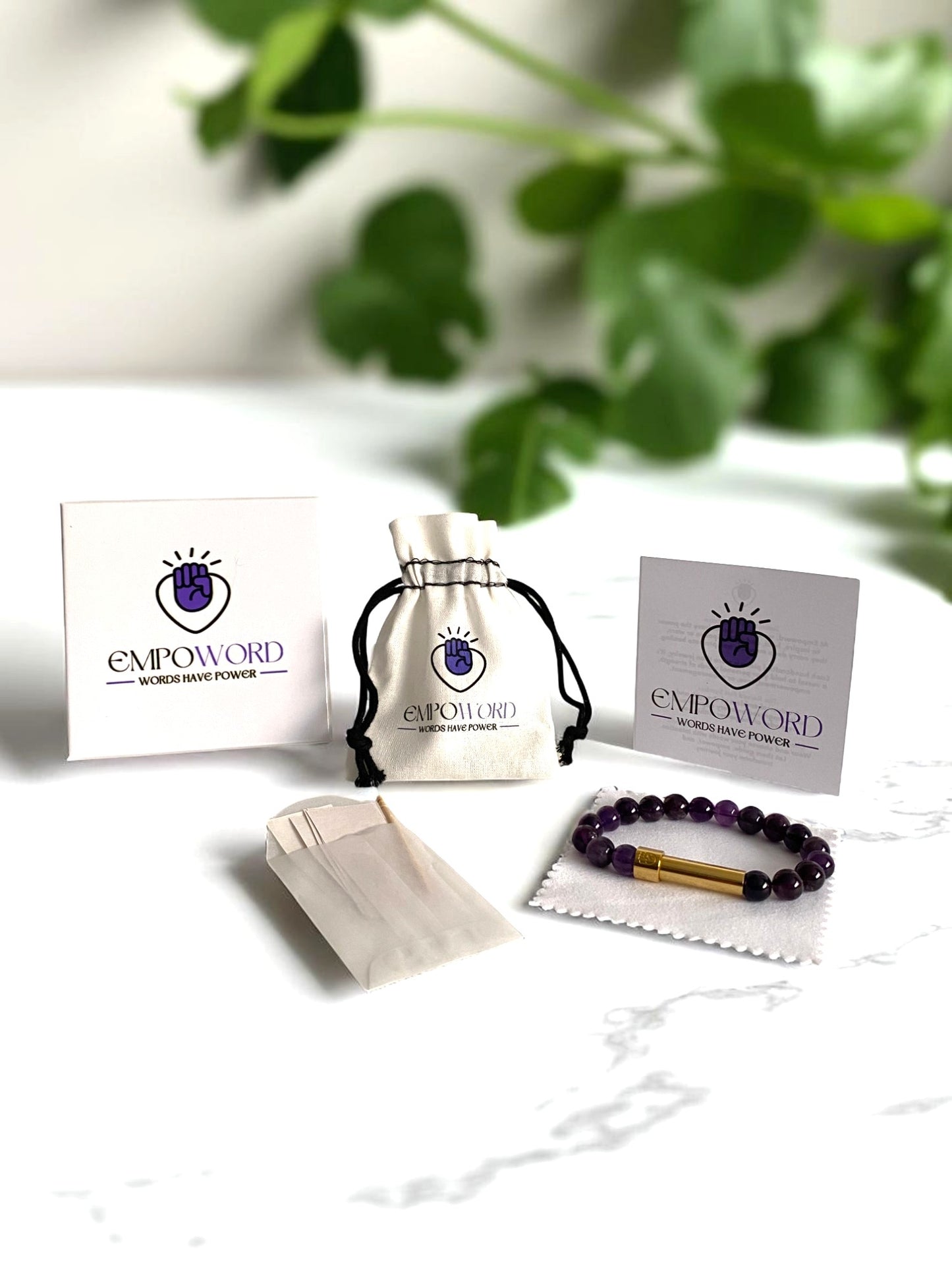 Empoword jewelry product packaging display featuring a purple beaded bracelet with a gold message capsule on a white cloth, a drawstring pouch with the Empoword logo, branded card and box with the slogan "Words Have Power," and a small paper envelope for handwritten notes, and white polishing cloth. Set on a white marble surface with green leaves in the blurred background, showcasing an elegant and empowering gift presentation.