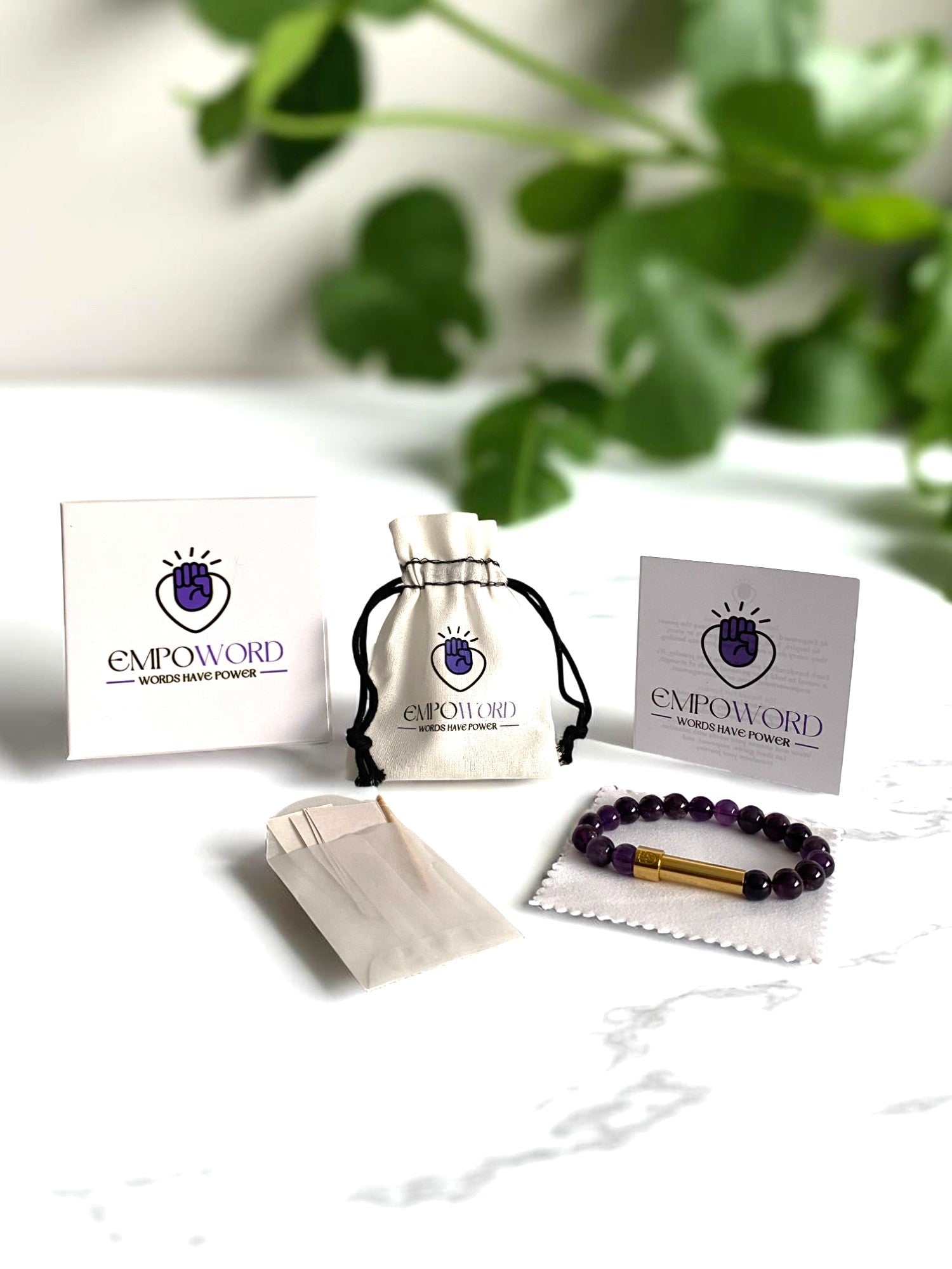 Empoword jewelry product packaging display featuring a purple beaded bracelet with a gold message capsule on a white cloth, a drawstring pouch with the Empoword logo, branded card and box with the slogan "Words Have Power," and a small paper envelope for handwritten notes, and white polishing cloth. Set on a white marble surface with green leaves in the blurred background, showcasing an elegant and empowering gift presentation.